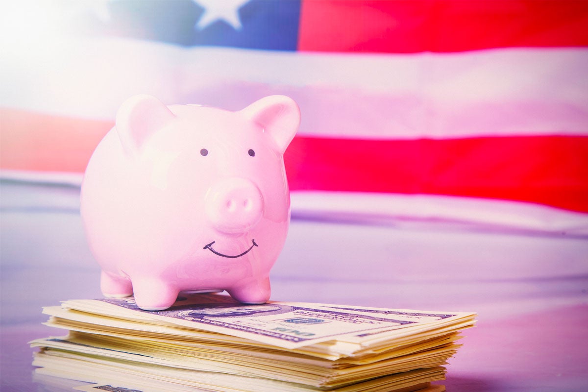 Piggy bank sitting on a pile of bills with an American flag in the background