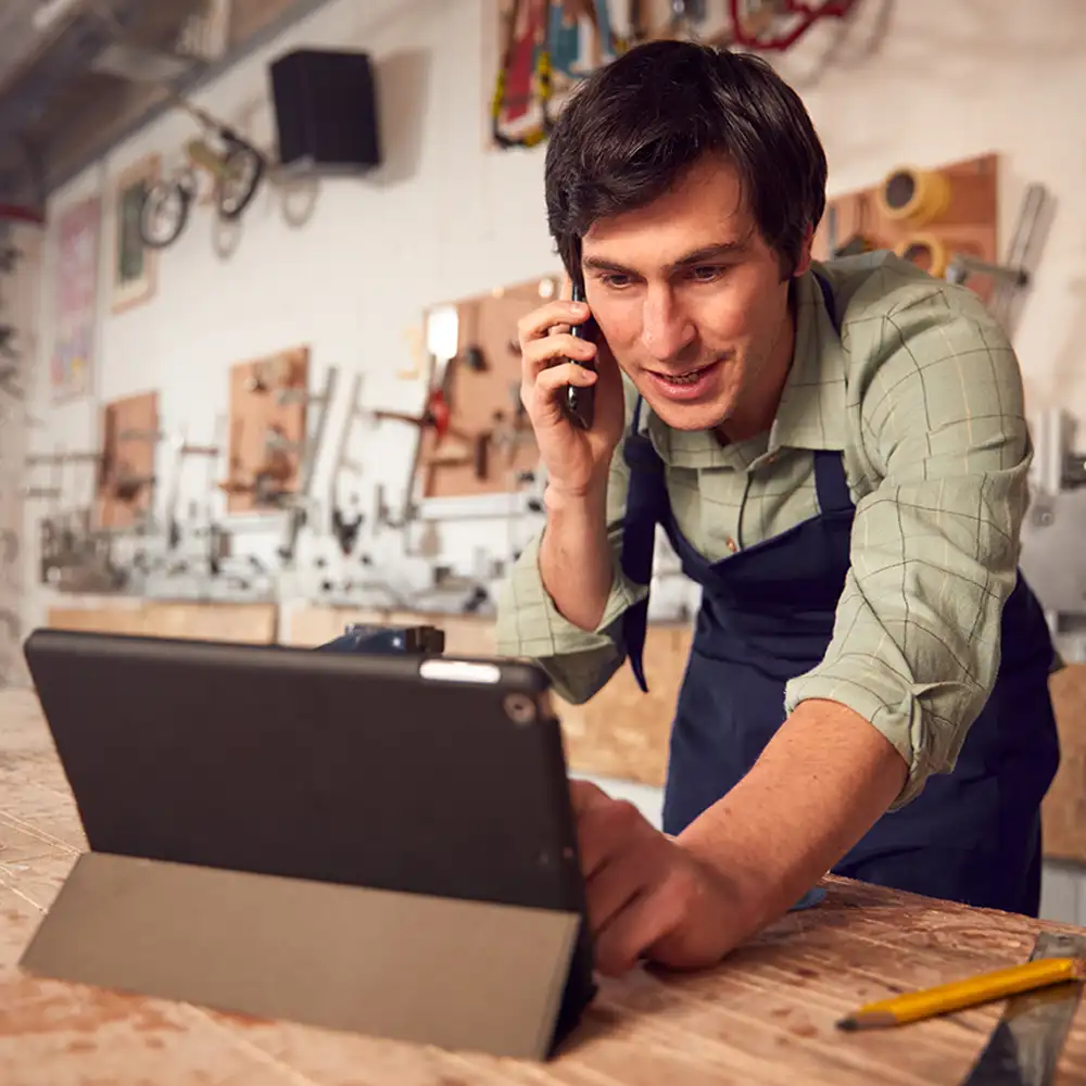 Man in workshop leaning down looking at a tablet computer while on a phone.